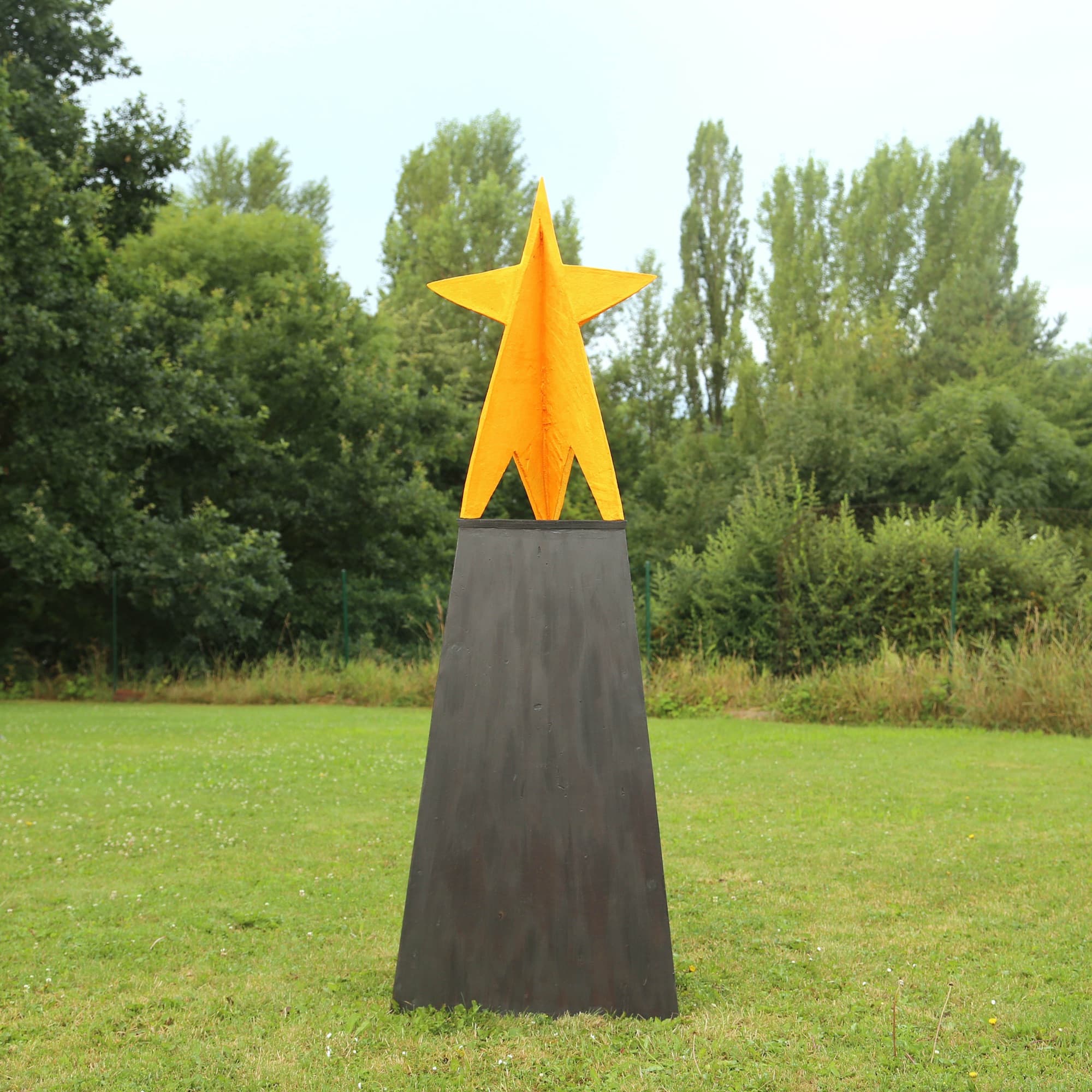Ester II, yellow star on a dark tapered plinth on a lawn, front view